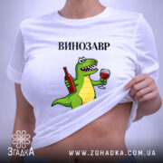 Футболка Винозавр, біла, на плоскій поверхні, з етикеткою, фон світло-сірий, принт з зеленим динозавром, що тримає пляшку вина та келих, текст 'ВИНОЗАВР' над принтом.