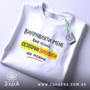 Футболка виправляти мене вже пізно, біла футболка, зображена зверху, фон світло-сірий, принт чорного, жовтого та червоного кольорів з текстом на українській мові, з етикеткою на комірці.