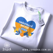 Футболка випускник 2026 року, біла, на столі, з принтом у формі серця, що містить жовті та блакитні кольори, напис 'НАЙКРАЩИЙ ВИПУСК 2026', деталі принта чітко видимі, фон світло-сірий.