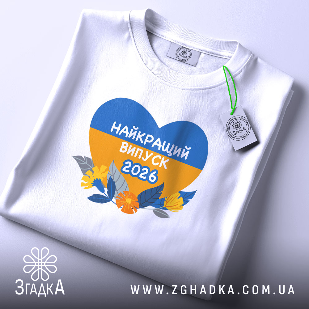 Футболка випускник 2026 року, біла, на столі, з принтом у формі серця, що містить жовті та блакитні кольори, напис 'НАЙКРАЩИЙ ВИПУСК 2026', деталі принта чітко видимі, фон світло-сірий.