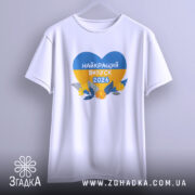 Футболка випускник 2026 року, біла, підвішена на вішалці, з принтом у формі серця, що містить жовті та блакитні кольори, напис 'НАЙКРАЩИЙ ВИПУСК 2026', фон світло-сірий.