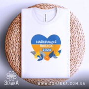 Футболка випускник 2026 року, біла, на круглому кошику, з принтом у формі серця, що містить жовті та блакитні кольори, напис 'НАЙКРАЩИЙ ВИПУСК 2026', фон світло-сірий.