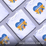 Футболка випускник 2026 року, біла, на моделі, з принтом у формі серця, що містить жовті та блакитні кольори, напис 'НАЙКРАЩИЙ ВИПУСК 2026', оточений квітами та листям, фон світло-фіолетовий.