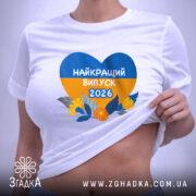 Футболка випускник 2026 року, біла, на столі, з принтом у формі серця, що містить жовті та блакитні кольори, напис 'НАЙКРАЩИЙ ВИПУСК 2026', деталі принта чітко видимі, фон світло-сірий.