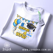 Футболка випускнику 4 класу, біла, на столі, з етикеткою, принт з написом 'Я вижив після 4 років НУШ', з ілюстраціями повітряних кульок і шапки випускника, яскраві кольори, святковий настрій.
