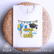 Футболка випускнику 4 класу, біла, складена на плетеній кошику, принт з написом 'Я вижив після 4 років НУШ', з ілюстраціями повітряних кульок і шапки випускника, яскраві кольори, святковий настрій.