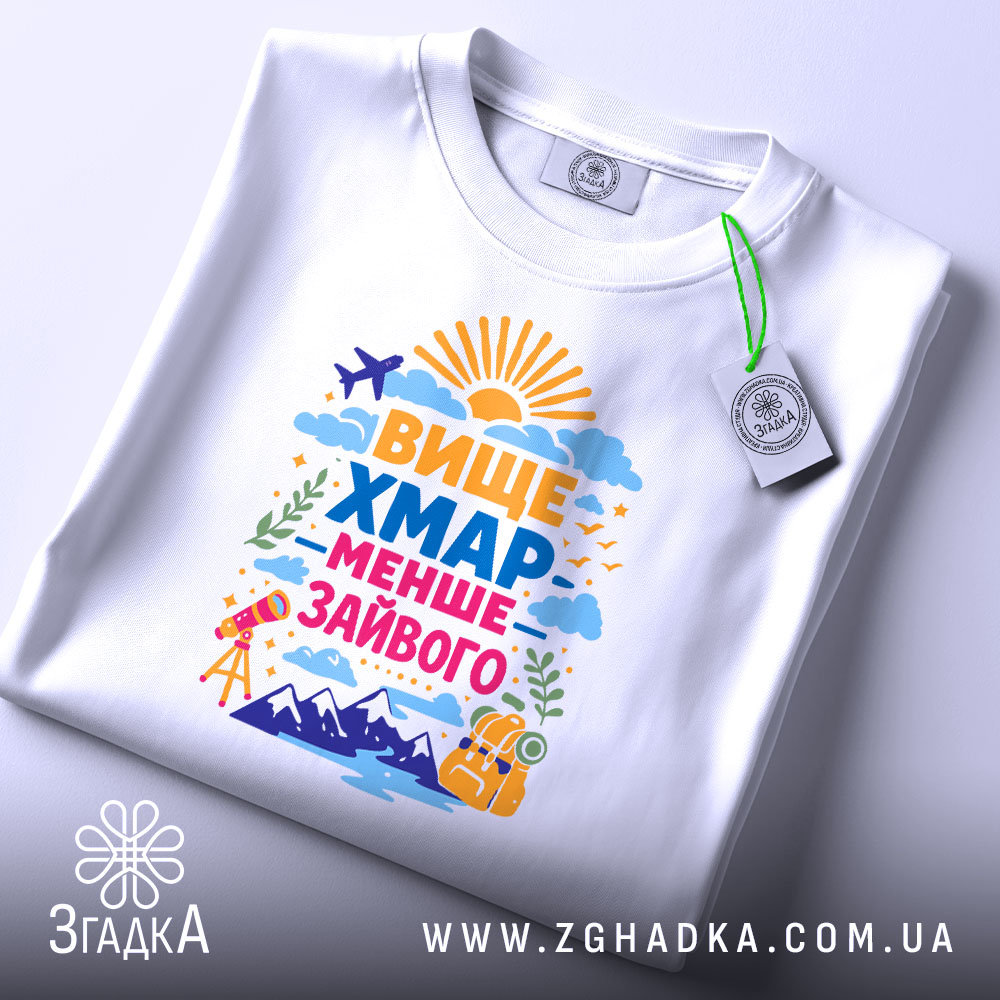 Футболка вище хмар менше зайвого, біла, на плоскій поверхні, з етикеткою, фон світлий, принт яскравий, з написом 'ВИЩЕ ХМАР МЕНШЕ ЗАЙВОГО' та малюнками сонця, літака, гір і телескопа.