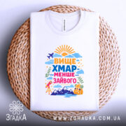 Футболка вище хмар менше зайвого, біла, на плоскій поверхні, з легким текстильним фоном, принт яскравий, з написом 'ВИЩЕ ХМАР МЕНШЕ ЗАЙВОГО' та малюнками сонця, літака, гір і телескопа.