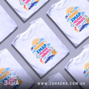 Футболка вище хмар менше зайвого, біла, на моделі, з легким макіяжем, фон пастельного кольору, принт з яскравими кольорами, з написом 'ВИЩЕ ХМАР МЕНШЕ ЗАЙВОГО' та ілюстраціями сонця, літака, гір та рюкзака.