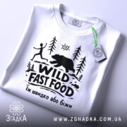 Футболка Wild Fast Food білого кольору, складена на поверхні, з видимим етикеткою, фон світлий, принт чорного кольору з малюнком людини, що біжить, та ведмедя на фоні лісу.