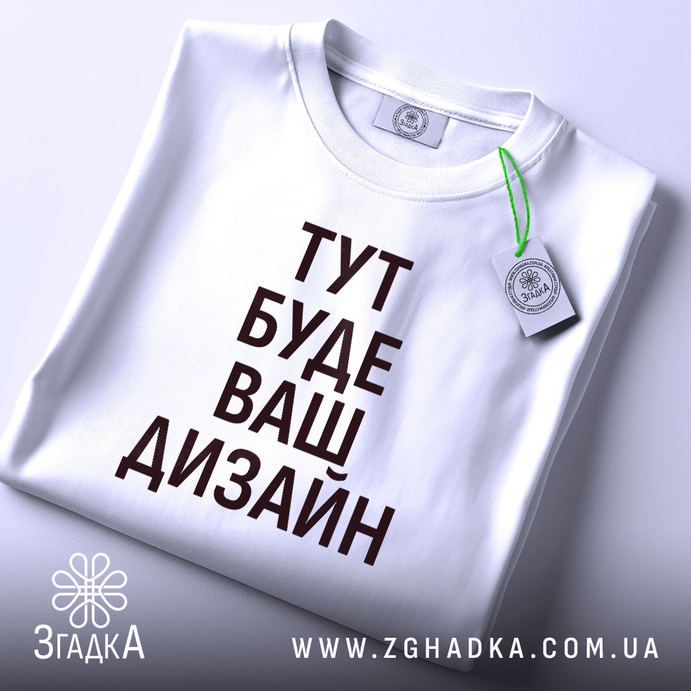 Футболка з вашим дизайном, білого кольору, зображена на столі в горизонтальному положенні, фон світло-фіолетовий, принт чорного кольору з текстом 'ТУТ БУДЕ ВАШ ДИЗАЙН', на комірці видна етикетка.