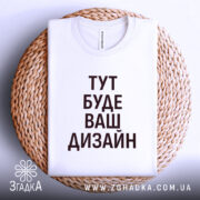 Футболка з вашим дизайном, білого кольору, складена на плетеній кошику, фон світло-фіолетовий, принт чорного кольору з текстом 'ТУТ БУДЕ ВАШ ДИЗАЙН'.