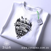 Футболка залишаю зайве внизу. Біла футболка, з принтом чорного кольору, розташована на плоскій поверхні. Принт містить зображення гір, лісу, сонця та різних предметів. На футболці є етикетка з зеленим шнурком. Фон світло-сірий.