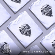 Футболка залишаю зайве внизу. Біла футболка на моделі, з коротким рукавом, з принтом чорного кольору, що зображає гори, ліс, сонце та різні предмети. Фон світло-фіолетовий, модель позує з легким усмішкою.