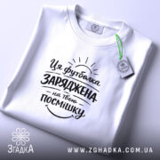 Футболка заряджена на твою посмішку. Біла футболка, складена на столі, з принтом чорного кольору, що містить текст і графічні елементи, з зеленим ярликом, на фоні світло-сірого кольору.