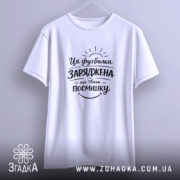 Футболка заряджена на твою посмішку. Біла футболка, підвішена на вішалці, з коротким рукавом, з принтом чорного кольору, що містить текст і графічні елементи, на фоні світло-сірого кольору.