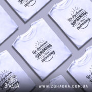 Футболка заряджена на твою посмішку. Біла футболка на чоловікові, з коротким рукавом, з принтом чорного кольору, що містить текст і графічні елементи, на фоні світло-фіолетового кольору.