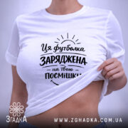 Футболка заряджена на твою посмішку. Біла футболка, складена на столі, з принтом чорного кольору, що містить текст і графічні елементи, з зеленим ярликом, на фоні світло-сірого кольору.