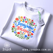 Футболка заряджена теплом, біла, на столі, з кольоровим принтом, що містить квіти та напис 'Заряджена теплом', фон світло-фіолетовий, деталі принта яскраві та чіткі.