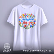 Футболка заряджена теплом, біла, підвішена на вішалці, з кольоровим принтом, що містить квіти та напис 'Заряджена теплом', фон світло-сірий, деталі принта яскраві та привабливі.