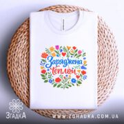 Футболка заряджена теплом, біла, на плетеній основі, з кольоровим принтом, що містить квіти та напис 'Заряджена теплом', фон світло-фіолетовий, деталі принта яскраві та гармонійні.