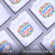 Футболка заряджена теплом, біла, на моделі, з кольоровим принтом, що містить квіти та напис 'Заряджена теплом', фон світло-фіолетовий, деталі принта яскраві та різноманітні.