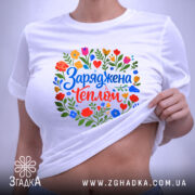 Футболка заряджена теплом, біла, на столі, з кольоровим принтом, що містить квіти та напис 'Заряджена теплом', фон світло-фіолетовий, деталі принта яскраві та чіткі.