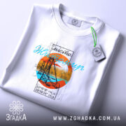 Футболка Житомир арт місто, біла, на столі, з легким ракурсом зверху, фон світлий, принт з оранжевим колом, синім текстом та зображенням мосту, етикетка на зеленій стрічці.