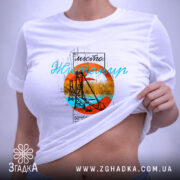 Футболка Житомир арт місто, біла, на столі, з легким ракурсом зверху, фон світлий, принт з оранжевим колом, синім текстом та зображенням мосту, етикетка на зеленій стрічці.