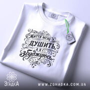 Футболка життя мене душить, біла футболка, складена на поверхні, фон світлий, принт чорного кольору з написом "Життя мене душить, а я збуджуюсь", етикетка з зеленим шнурком.