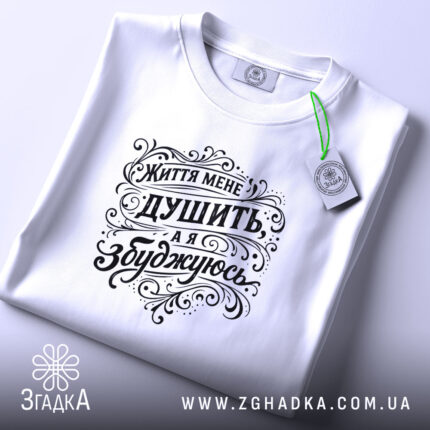 Футболка життя мене душить, біла футболка, складена на поверхні, фон світлий, принт чорного кольору з написом "Життя мене душить, а я збуджуюсь", етикетка з зеленим шнурком.