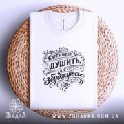 Футболка життя мене душить, біла футболка, розкладена на плетеній кошику, фон світлий, принт чорного кольору з написом "Життя мене душить, а я збуджуюсь", вид зверху.