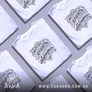 Футболка життя мене душить, біла футболка з чорним принтом, зображена з ракурсу спереду, фон світло-фіолетовий, принт містить напис "Життя мене душить, а я збуджуюсь" у декоративному стилі.