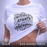 Футболка життя мене душить, біла футболка, складена на поверхні, фон світлий, принт чорного кольору з написом "Життя мене душить, а я збуджуюсь", етикетка з зеленим шнурком.