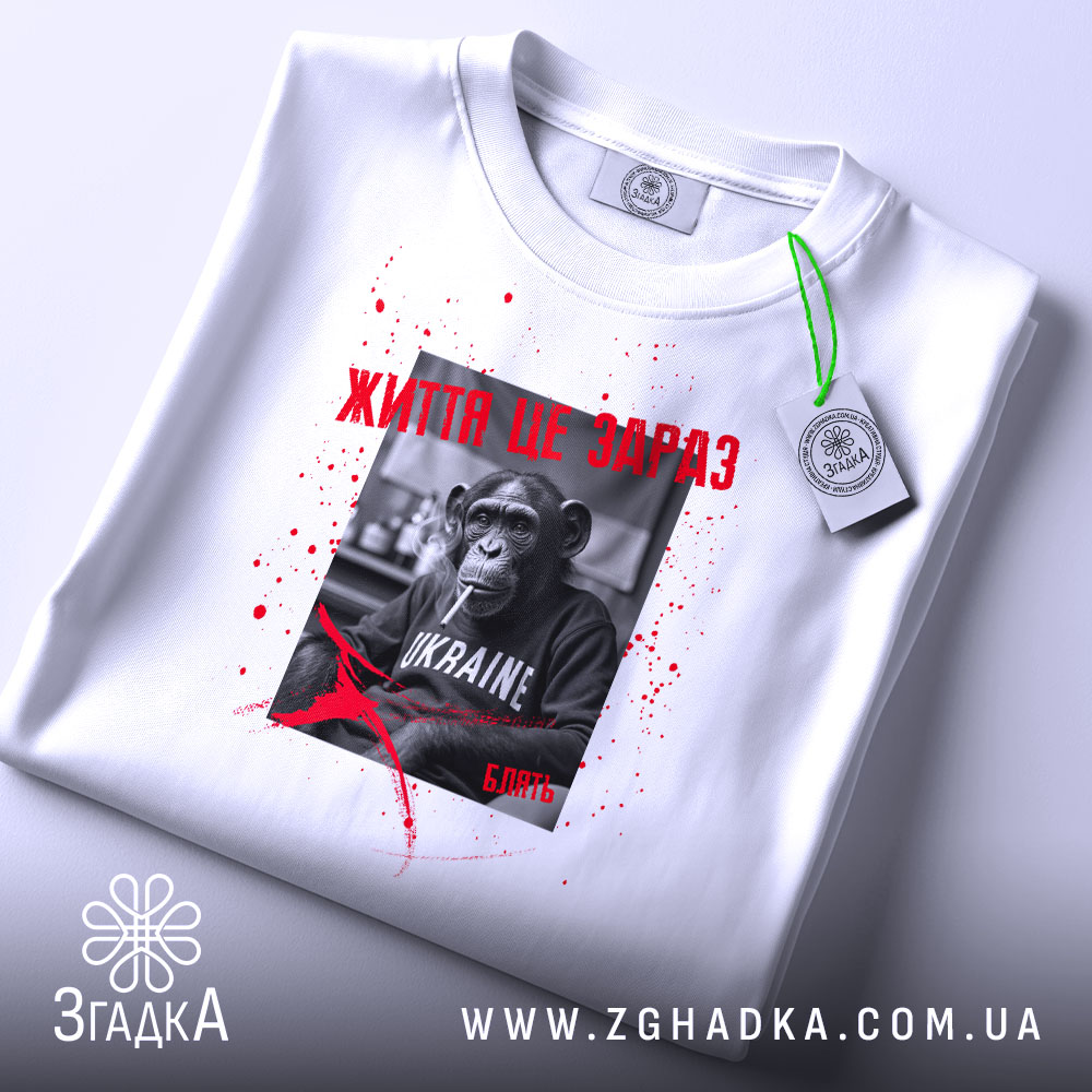 Футболка Життя це зараз, біла футболка, зображена зверху, фон світло-фіолетовий, принт з чорно-білим шимпанзе, текстом червоного кольору та плямами червоного кольору, на ярлику зелена стрічка.