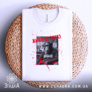 Футболка Життя це зараз, біла футболка, зображена зверху на плетеній кошику, фон білий, принт з чорно-білим шимпанзе, текстом червоного кольору та плямами червоного кольору.