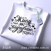 Футболка Живу як відчуваю, біла, складена на поверхні, фон світло-сірий, принт чорний з написом 'Живу як відчуваю' та декоративними елементами, з етикеткою зеленого кольору.