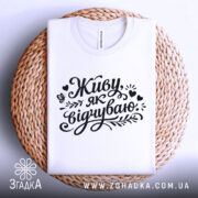 Футболка Живу як відчуваю, біла, складена на плетеній основі, фон світло-сірий, принт чорний з написом 'Живу як відчуваю' та декоративними елементами.