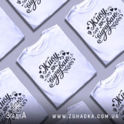 Футболка Живу як відчуваю, біла, зображена на моделі з коротким рукавом, фон світло-фіолетовий, принт чорний з написом 'Живу як відчуваю' та декоративними елементами.