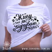 Футболка Живу як відчуваю, біла, складена на поверхні, фон світло-сірий, принт чорний з написом 'Живу як відчуваю' та декоративними елементами, з етикеткою зеленого кольору.