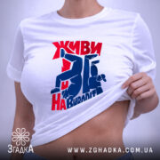 Футболка Живи навиворіт, біла, на столі, з принтом червоного та синього кольору, фон світло-сірий, принт з написом та фігурами людей у динамічних позах, з етикеткою на шнурку.