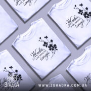 Футболка живи не існуй, біла, зображена з близького ракурсу, на фоні світло-сірого кольору, з чорним принтом, що містить напис 'Живи, а не просто існуй' та квітковий візерунок.