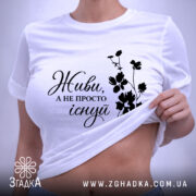 Футболка живи не існуй, біла, зображена зверху, на фоні світло-сірого кольору, з чорним принтом, що містить напис 'Живи, а не просто існуй' та квітковий візерунок, з етикеткою на зеленій стрічці.