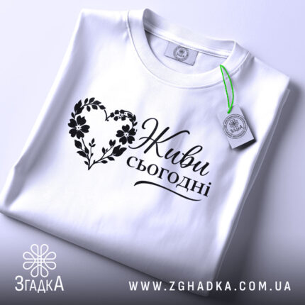 Футболка живи сьогодні, біла, складена на поверхні, ракурс зверху, фон світло-фіолетовий, принт чорного кольору з написом 'Живи сьогодні' у формі серця з квітами, з етикеткою зеленого кольору.