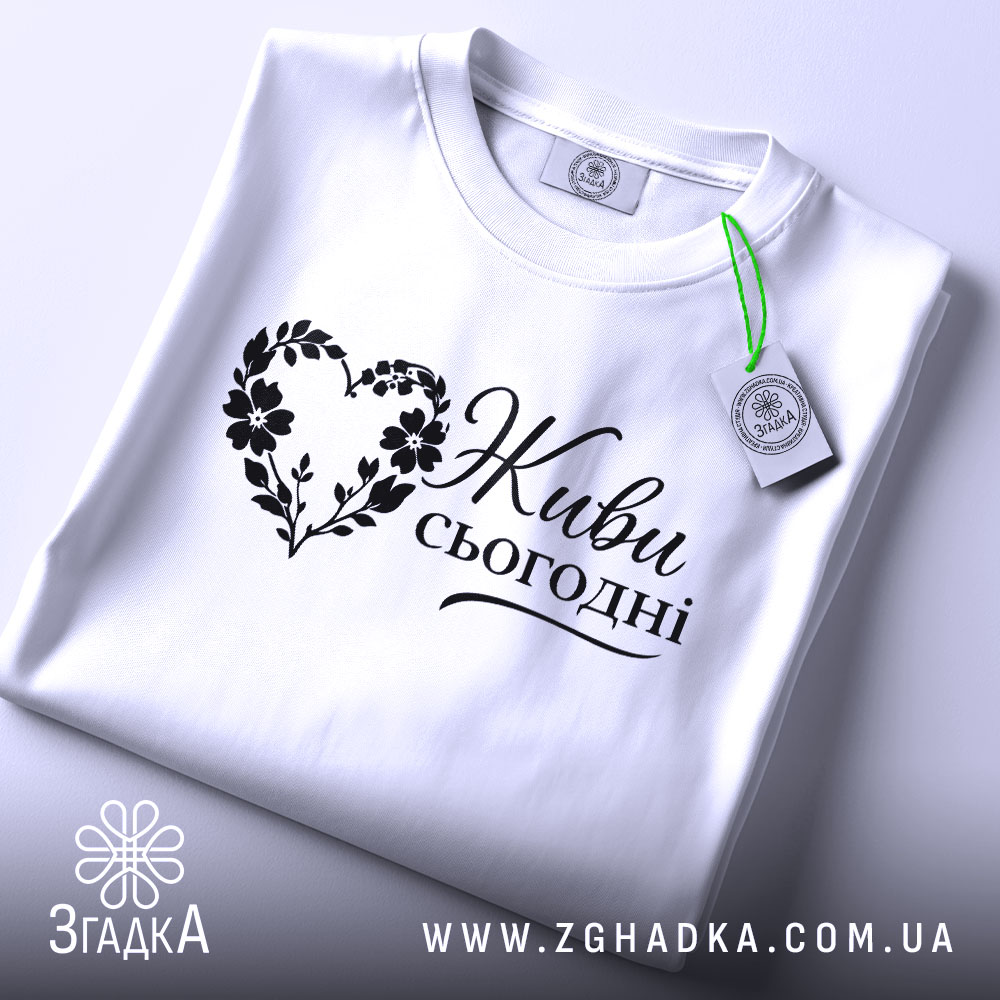 Футболка живи сьогодні, біла, складена на поверхні, ракурс зверху, фон світло-фіолетовий, принт чорного кольору з написом 'Живи сьогодні' у формі серця з квітами, з етикеткою зеленого кольору.
