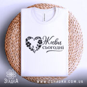 Футболка живи сьогодні, біла, складена на кошику, ракурс зверху, фон світло-фіолетовий, принт чорного кольору з написом 'Живи сьогодні' у формі серця з квітами.