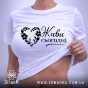 Футболка живи сьогодні, біла, складена на поверхні, ракурс зверху, фон світло-фіолетовий, принт чорного кольору з написом 'Живи сьогодні' у формі серця з квітами, з етикеткою зеленого кольору.