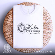 Футболка Живи тут тепер білого кольору, складена на плетеній кошику. Принт з годинником та написом виконаний у чорному кольорі. Фон світлий, що надає зображенню затишного вигляду.