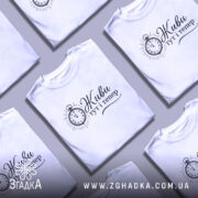 Футболка Живи тут тепер білого кольору, зображена на молодій людині, яка позує з легким усмішкою. Принт на футболці містить годинник та напис, виконаний у чорному кольорі. Фон світло-фіолетовий, що підкреслює яскравість футболки.