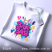 Футболка зла зая, білого кольору, розкладена на поверхні, фон світло-сірий, принт яскравий, з зображенням злого зайця та кольоровими сплесками фарби, етикетка з зеленим шнурком.